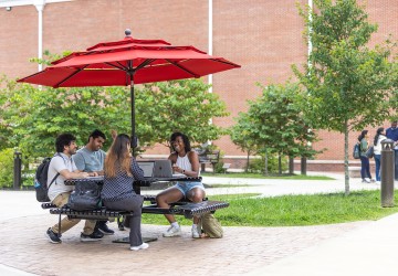 Four students sit at a table with their computers studying outside. 