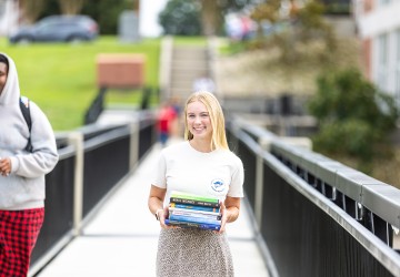 A female student stands on the campus viaduct holding a stack of textbooks. 