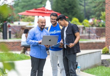 Three male students, one in a Cumberlands shirt, look at a laptop together in the campus quad. 