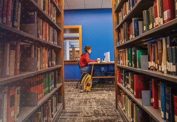 A female student sits at a booth in the library studying with shelves of books in the foreground.