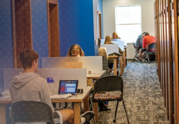 Several students sit at study desks inside the campus library with book shelves to their right. 