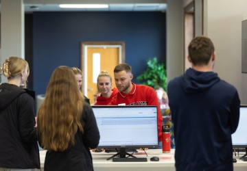 A group of students watch a male student demonstrate using AI during an info session in the student center. 