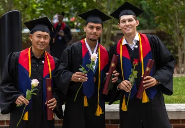 Three male students hold a rose and their diploma following graduation. 