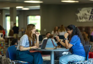 Two female students sit with their laptops at a round table in the student center. 