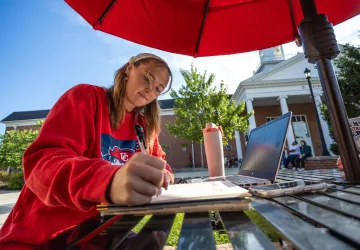 A female student sits at a table outside on campus and writes in her notebook. 