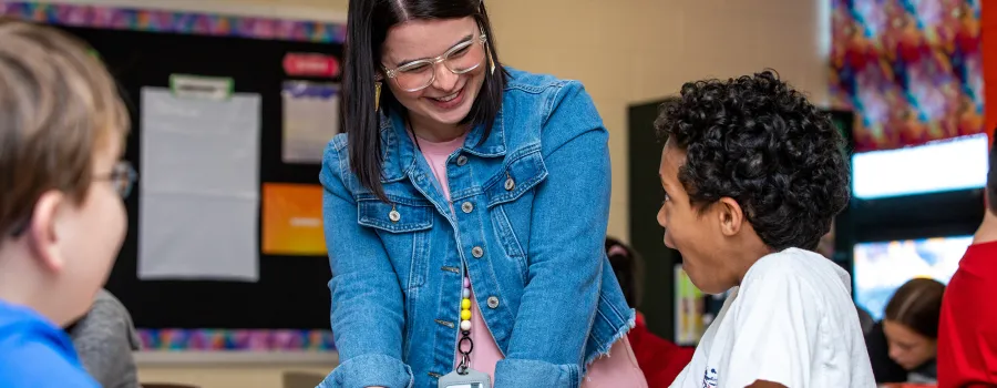  A teacher talking to students sitting at a desk while in a classroom. 