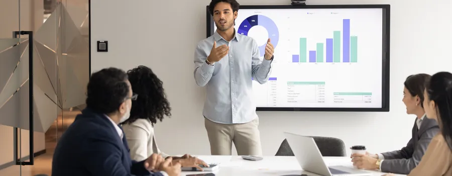 A young man shows data on a computer screen to a team of four professionals sitting at a table. 