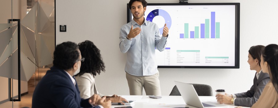 A young man shows data on a computer screen to a team of four professionals sitting at a table. 