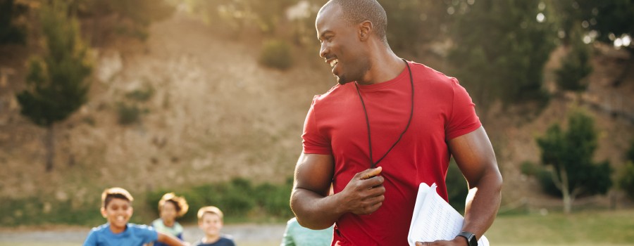A male P.E. teacher leads children in a run outside. 