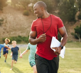 A male P.E. teacher leads children in a run outside. 