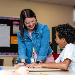  A teacher talking to students sitting at a desk while in a classroom. 