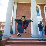 A graduate leaping for joy in front of a building. 