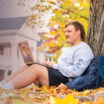 A student studies under a tree amongst fallen leaves on campus.
