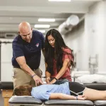 A physical therapy student is participating in a clinical exercise checking the back of another student with an instructor watching. 