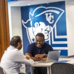 Two male students chat at a table with their computer inside the Patriot Success Center. 