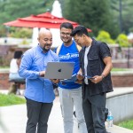 Three male students look at their computer while standing in the campus quad. 
