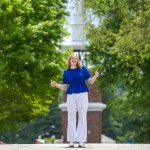 A female student in a blue shirt gives the thumbs up in front of the campus clock tower. 