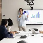 A young man shows data on a computer screen to a team of four professionals sitting at a table. 