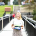A female student stands on the campus viaduct holding a stack of textbooks. 