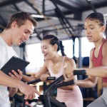 A male fitness instructor monitors a female working out on an exercise bike. 