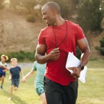 A male P.E. teacher leads children in a run outside. 