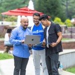 Three male students, one in a Cumberlands shirt, look at a laptop together in the campus quad. 