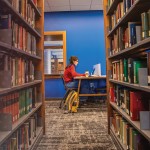 A female student sits at a booth in the library studying with shelves of books in the foreground.