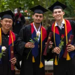 Three male students hold a rose and their diploma following graduation. 