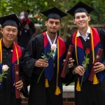 Three male students hold a rose and their diploma following graduation. 