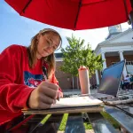 A female student sits at a table outside on campus and writes in her notebook. 