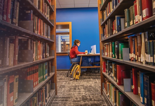 A female student sits at a booth in the library studying with shelves of books in the foreground.